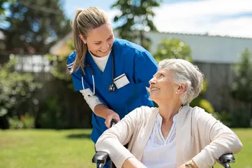 Jobway Germany - Internationale Rekrutierung von Fachkräften - Eine lächelnde Pflegekraft in blauer Arbeitskleidung schiebt eine ältere Frau im Rollstuhl durch einen sonnigen Garten. Die Frau lächelt ebenfalls und blickt zu der Pflegekraft auf.