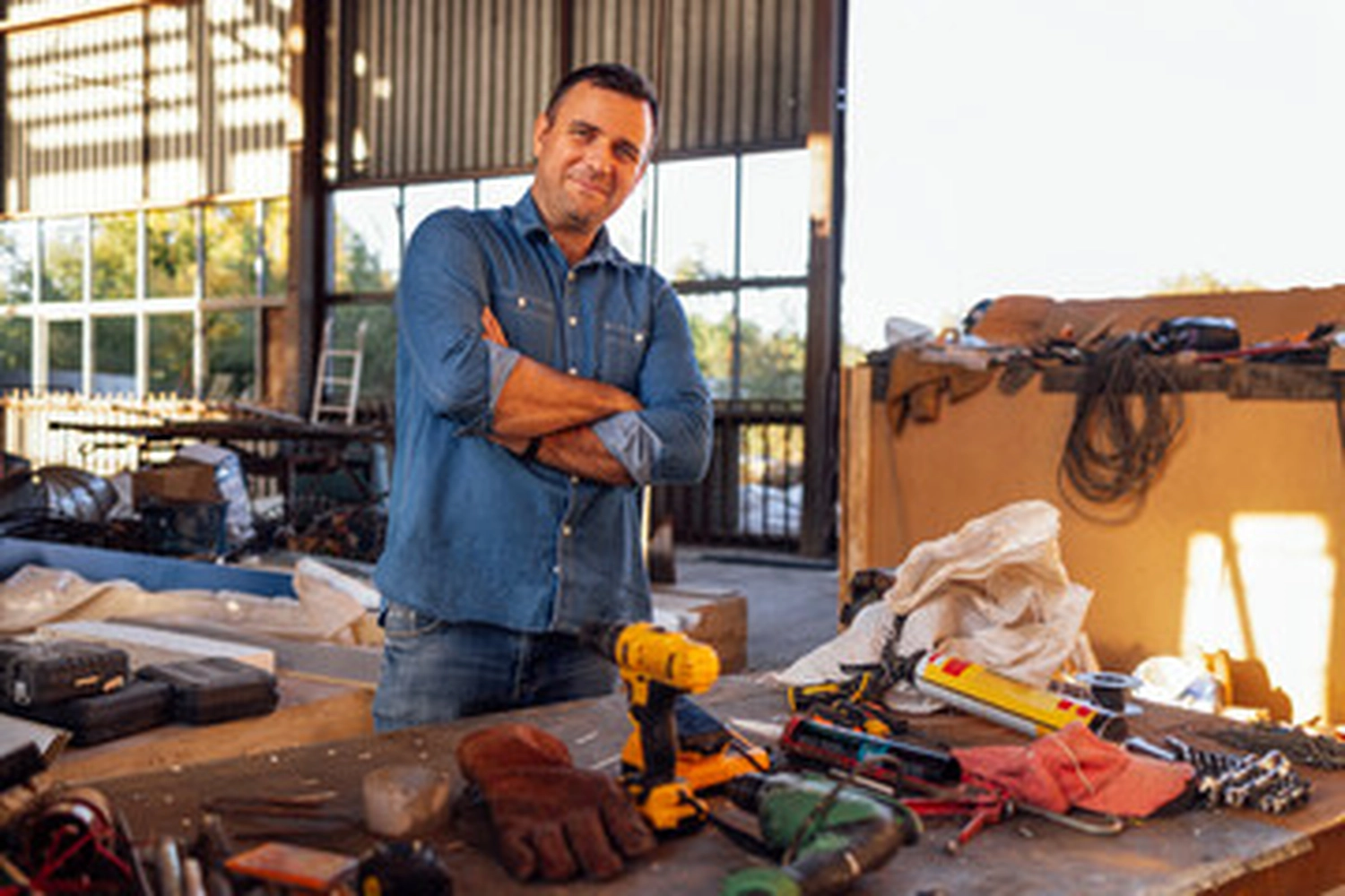 Jobway Germany - Internationale Rekrutierung von Fachkräften - Ein lächelnder Mann mittleren Alters in Jeanshemd und Jeans steht mit verschränkten Armen hinter einer Werkbank mit verschiedenen Elektrowerkzeugen und Handschuhen in einer Werkstatt.